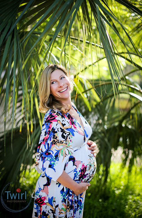 A pregnant woman wearing a floral maxi dress in front of palm leaves in the park of New Orleans. 
