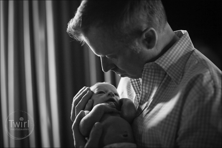 A father looking down at his newborn son near a window. 