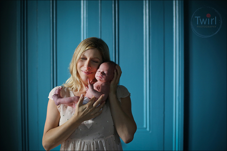 A mom holding a newborn up to her cheek and smiling in front of a blue wall at their home in New Orleans.