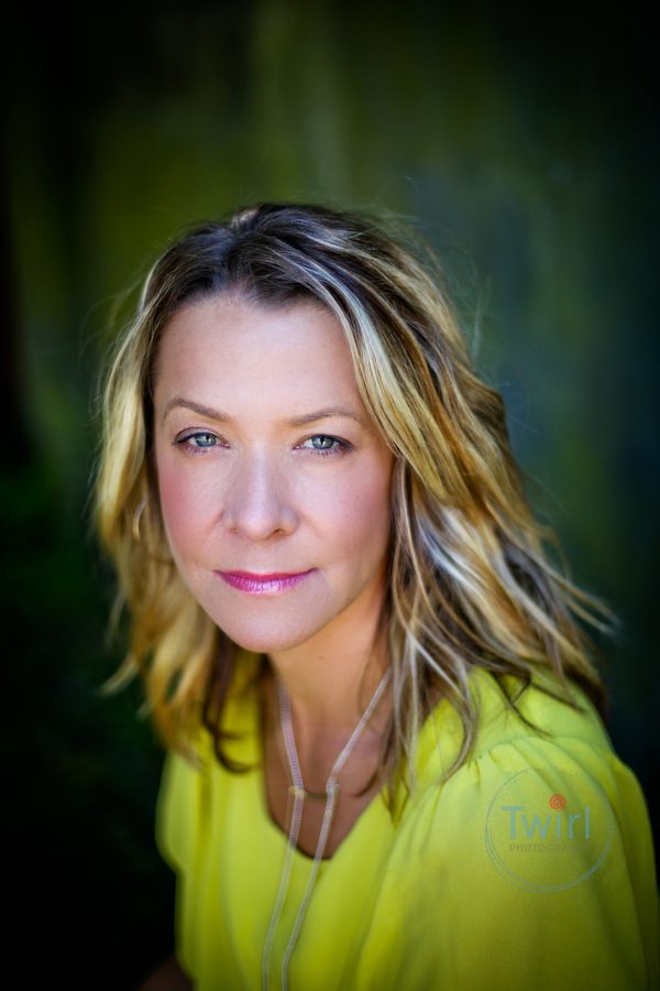 A headshot of a woman with blonde hair and a yellow shirt in front of a green and yellow wall in the Garden District for professional portraits.