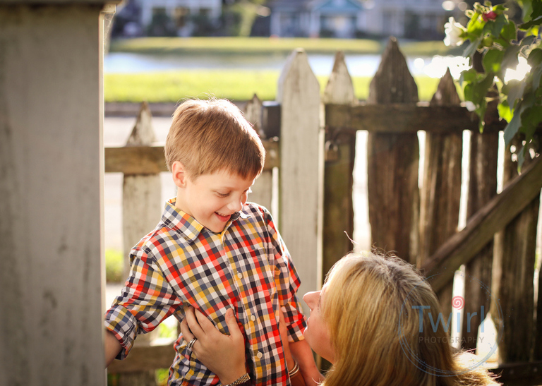 A mother looking up at her son in front of the bayou and a wood fence at the Pitot House for family photography.