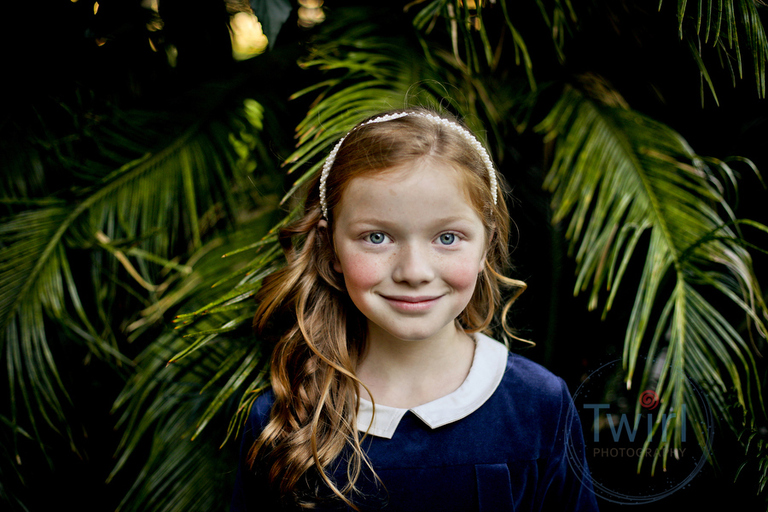 A girl with blue eyes and red hair wearing a blue dress with a peter pan collar standing in front of big palm leaves outside of the Pitot House for New Orleans Family Portraits