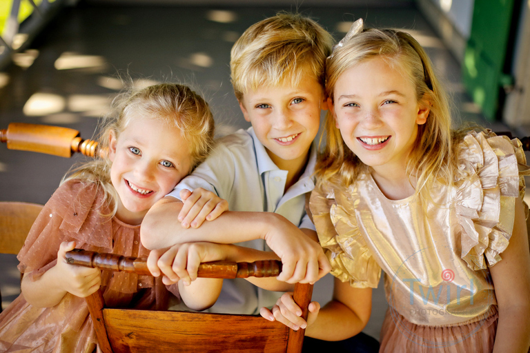 Three siblings laughing while siting on a wood chair wearing gold and rose gold dresses and a blue shirt on the balcony of the Pitot House in New Orleans for family photos.
