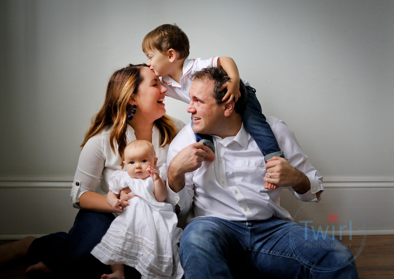A family sitting on the floor with big brother on dad's shoulders as he leans over to kiss mom's forehead while holding baby sister during newborn photos in New Orelans.