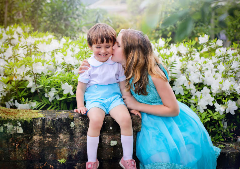 A brother and sister sitting near blooming Azalea flowers wearing turquoise blue dress and shorts for Spring photos at Longue Vue House and Gardens.