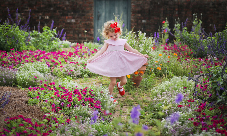 A little girl wearing a pink spring dress and red bow with matching mary jane shoes in the middle of flowers at the New Orleans Botanical Gardens for Spring Family Photography