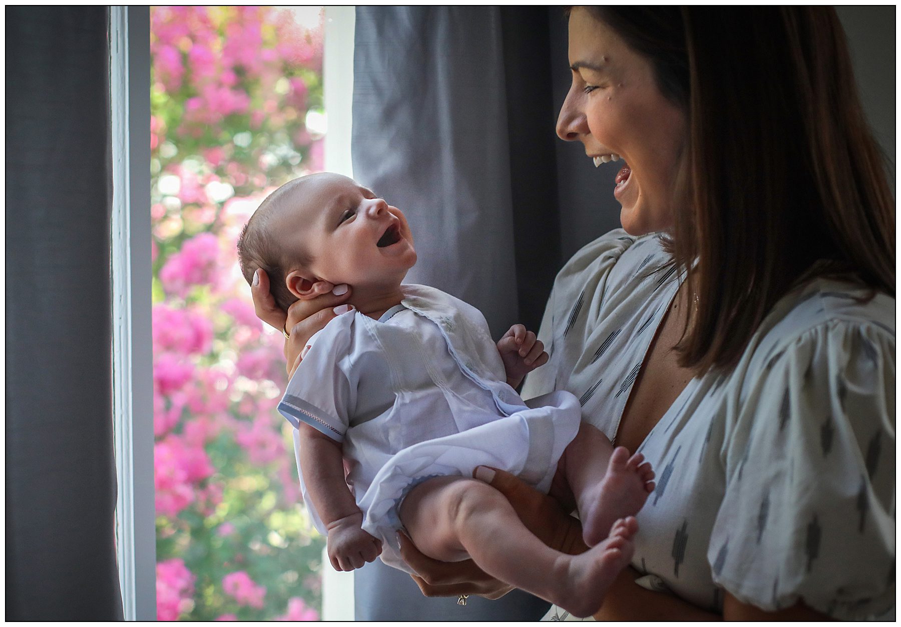 A 3 month old baby smiling at his mother as she holds him by a window with flowers outside during a newborn photography session in New Orleans.