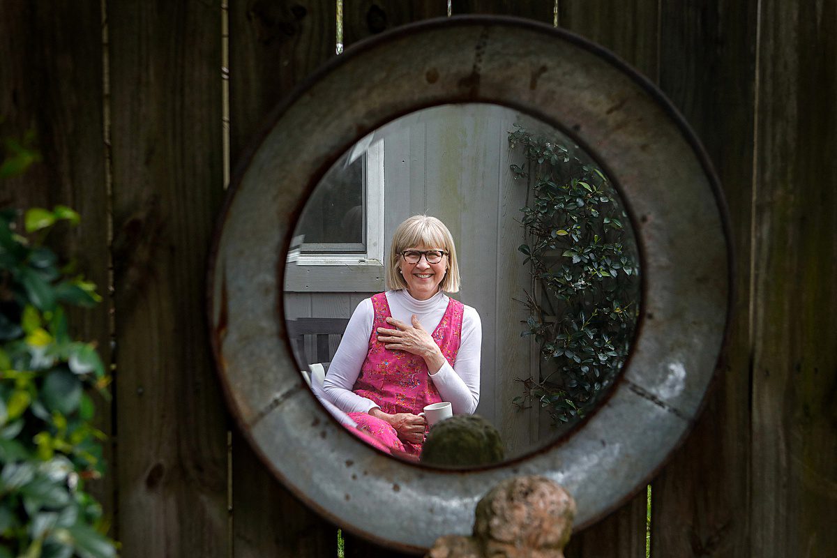 A woman reflected in a round mirror wearing a pink dress and smiling at the camera as she gets headshot photos taken.