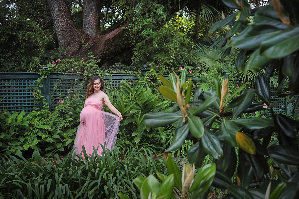 Outdoor maternity session at Longue Vue gardens in New Orleans with pregnant woman wearing pink dress standing in a lush green garden for New Orleans maternity photographer. Check out these date ideas in New Orleans before baby arrives.