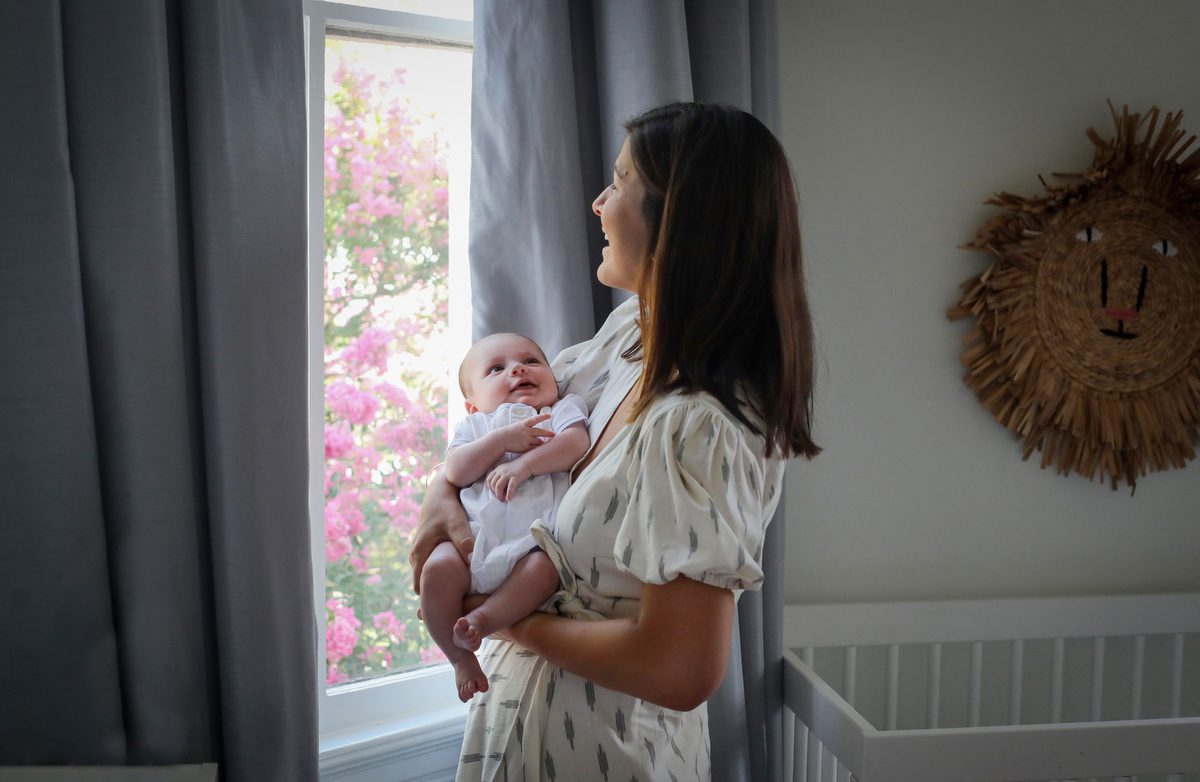 Mom wearing a stylish cream wrap dress holding her newborn son in his New Orleans home nursery during a lifestyle newborn photo session by Twirl Photography for an article about how to choose outfits for your lifestyle newborn photos.