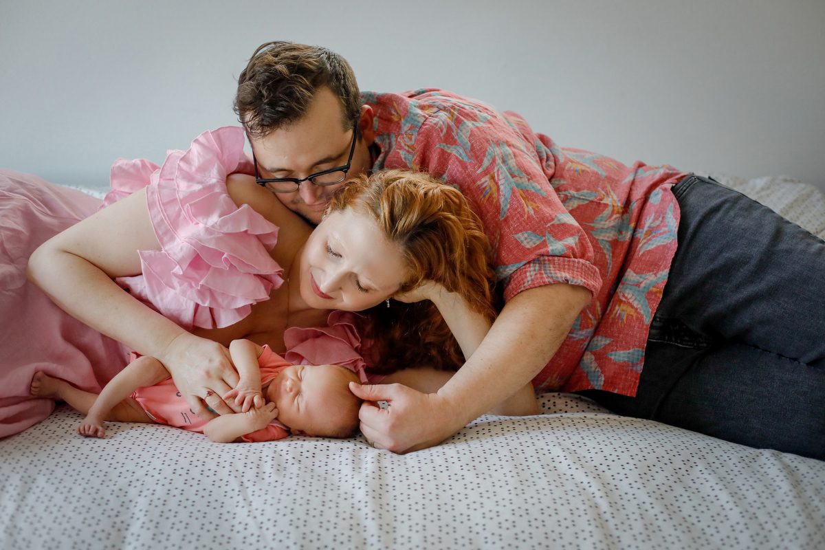 Dad and mom laying on a bed holding their newborn daughter during a lifestyle newborn photography session in New Orleans by Twirl Photography.