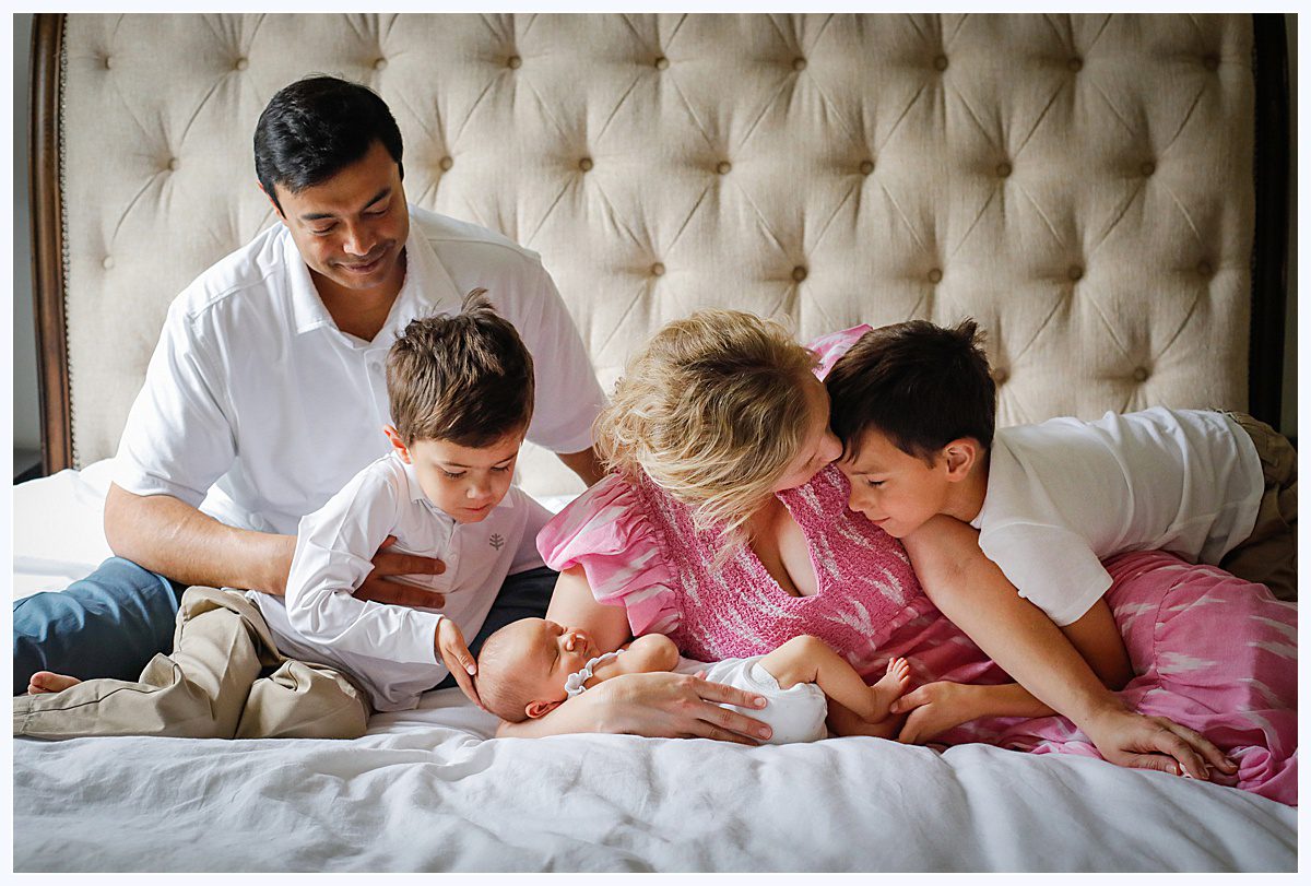 A family of mom, dad and two sons hug on a bed as they hold their newborn daughter during photos for New Orleans newborn photographer.