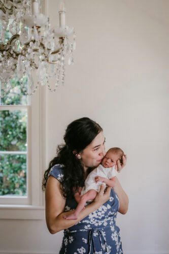 A mom leans down and kisses her newborn baby wearing a blue dress in her dining room.