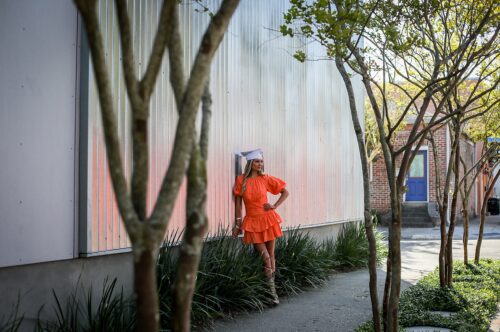 A senior in an orange dress leaning against a reflective house in between trees.