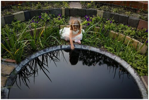 A little girl in a white dress reaching her hand into a round pond reflection the sky for a story about why I love to be a New Orleans Photographer.