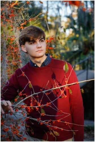 A high school senior posing for a portrait in a red sweater surrounded by orange foliage in the park.