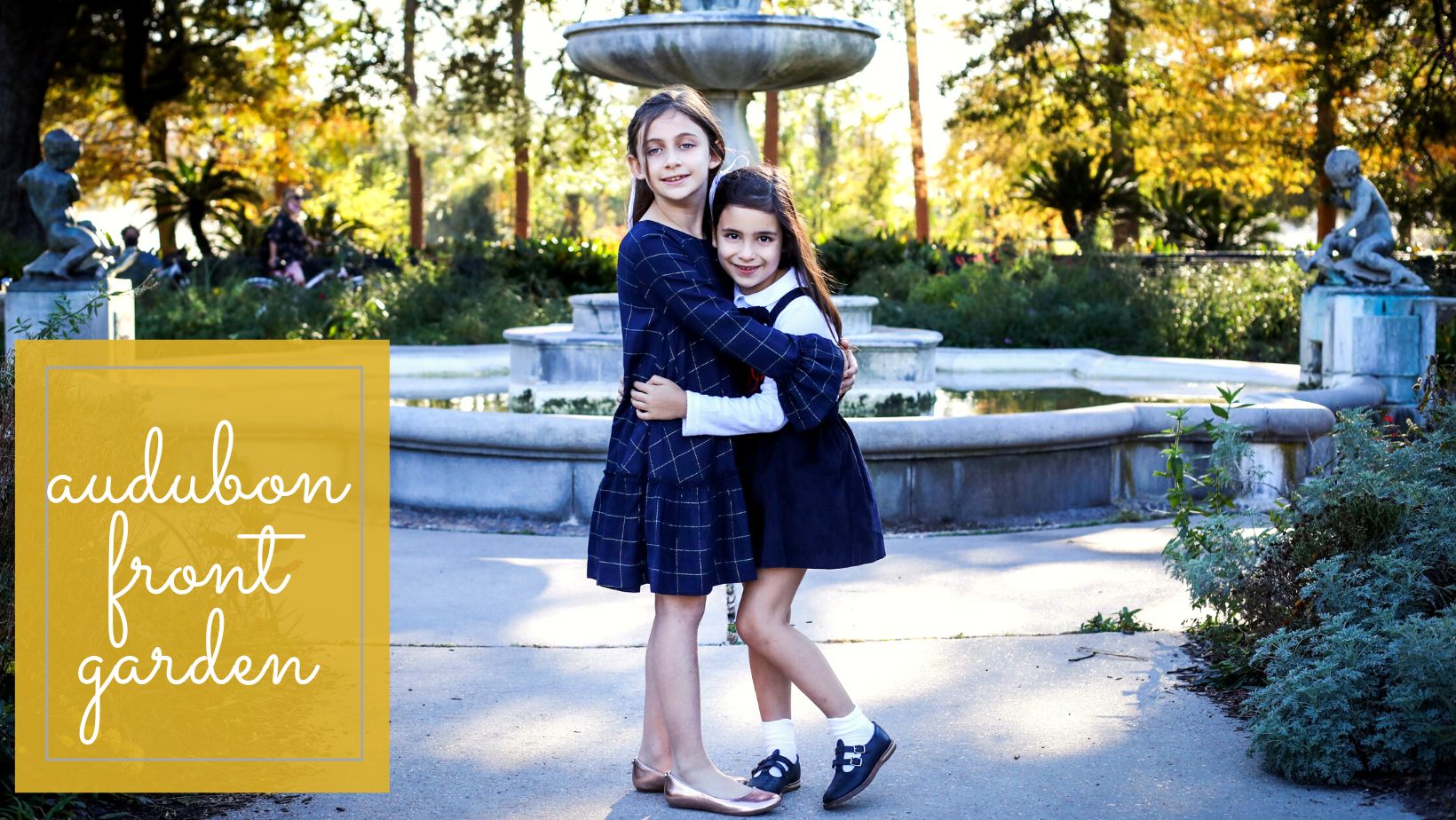 Two sisters hugging in front of a park fountain.