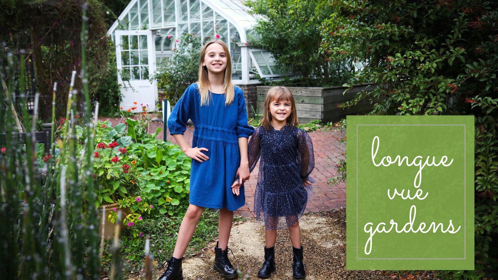 Two girls standing in a garden in front of a white greenhouse building.