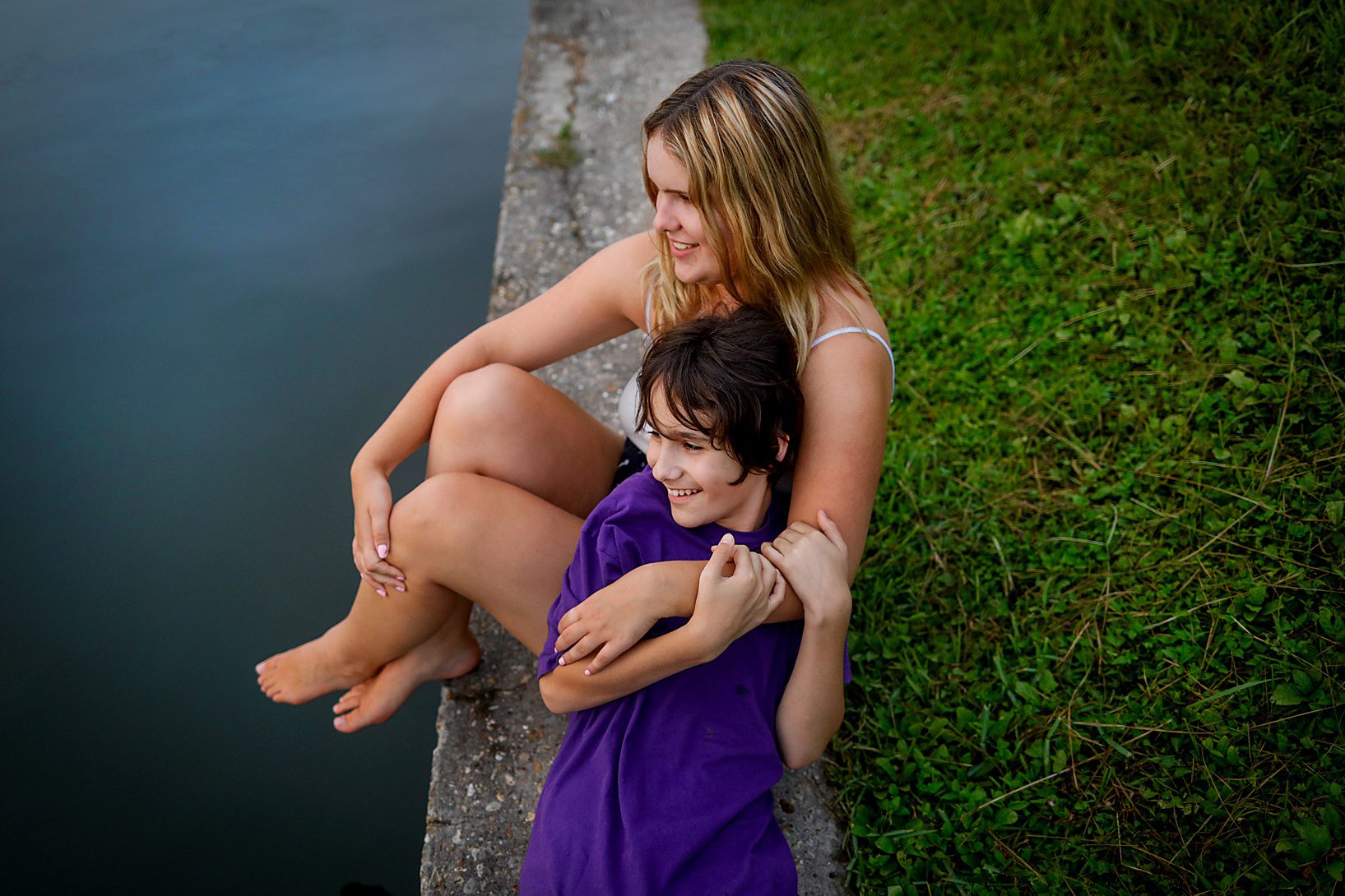 Two kids sitting on the edge of a bayou smiling.