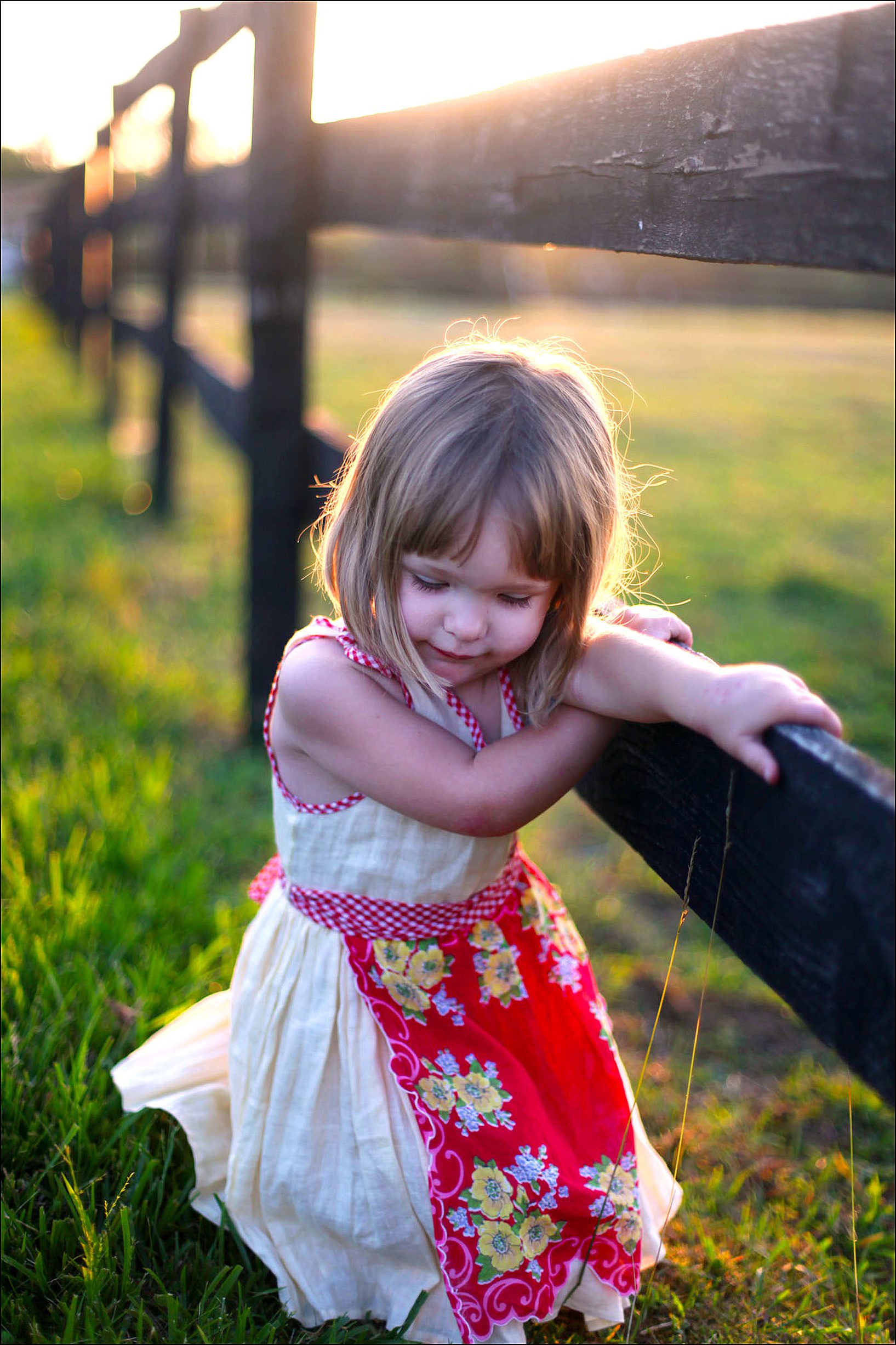 A girl wearing a yellow dress and red apron looking down while leaning on an old fence.