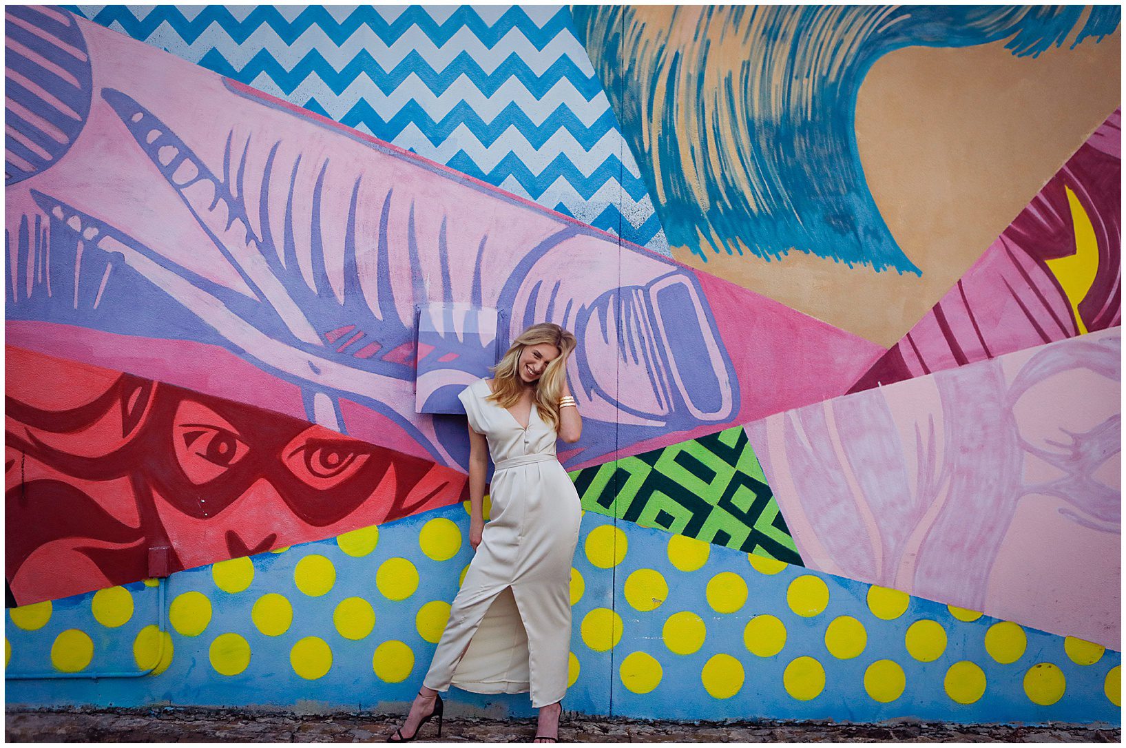 A high school senior in a long white dress standing in front of a colorful mural.