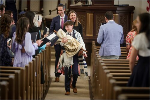 A young man walks the torah up the aisle at Touro Synagogue with his parents behind him.