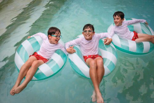 Three brothers with red bathing suits and white shirts sitting on green and white striped blow up floats in a pool.