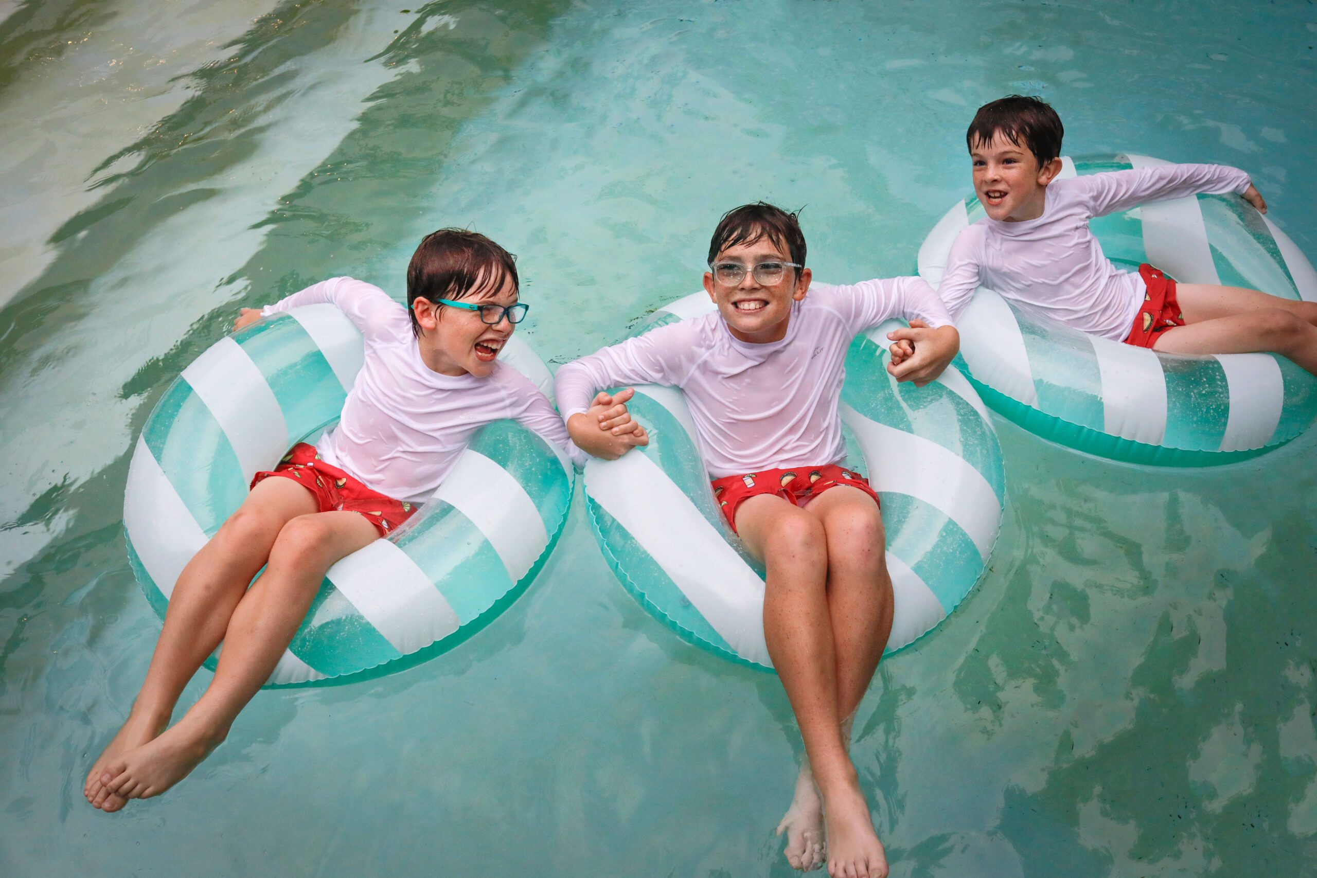 Three brothers with red bathing suits and white shirts sitting on green and white striped blow up floats in a pool.